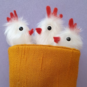 May include: Three white fluffy chicks with red combs and black eyes peeking out of a yellow fabric basket.