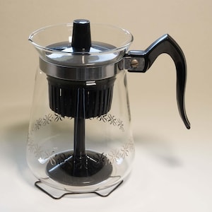 May include: A vintage glass coffee percolator with a black handle, lid, and internal components. The clear glass carafe features a decorative floral pattern and sits on a wire stand. A classic kitchen appliance.