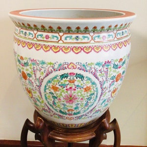May include: A white ceramic planter with a floral design in shades of pink, green, blue, and yellow. The planter is on a wooden stand.