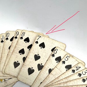 May include: A fan of antique playing cards featuring the suit of spades. The cards have a distressed, vintage appearance with black suit symbols and numbers. The seven of spades is prominently displayed, with other cards visible.