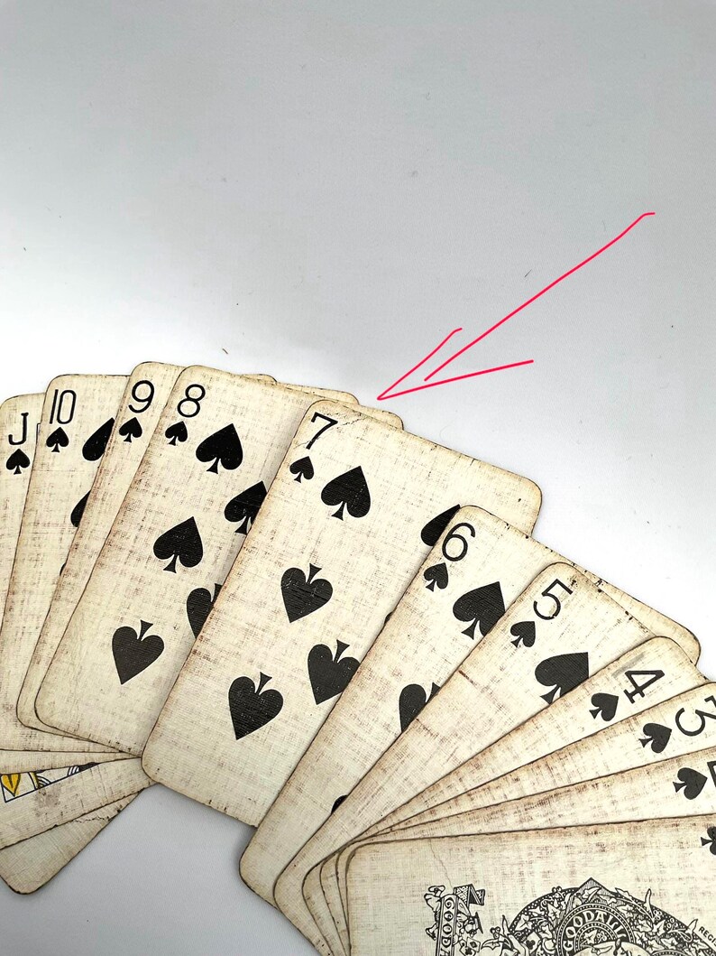 May include: A fan of antique playing cards featuring the suit of spades. The cards have a distressed, vintage appearance with black suit symbols and numbers. The seven of spades is prominently displayed, with other cards visible.