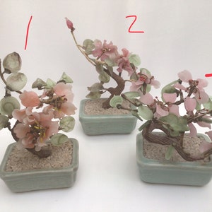 May include: Three miniature glass bonsai trees with pink flowers and green leaves in green ceramic pots. The trees are arranged in a row on a white surface.