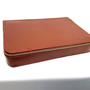 May include: A rectangular, brown leather case with a gold zip. The case has red stitching along the edges and appears slightly worn. The case is closed and sits on a white surface.