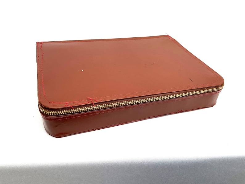 May include: A rectangular, brown leather case with a gold zip. The case has red stitching along the edges and appears slightly worn. The case is closed and sits on a white surface.