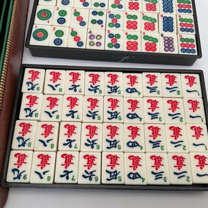 May include: A complete Mahjong set with ivory-coloured tiles featuring red, green, blue, and black symbols and characters. The tiles are arranged in black trays, with a green and brown zipped case visible on the left.