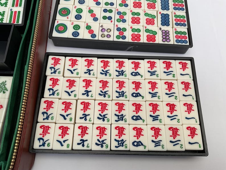 May include: A complete Mahjong set with ivory-coloured tiles featuring red, green, blue, and black symbols and characters. The tiles are arranged in black trays, with a green and brown zipped case visible on the left.