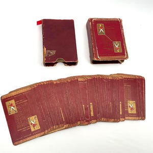 May include: Vintage playing cards with a burgundy and gold design. The deck is fanned out, revealing the card backs and a portion of the faces. The card case is also burgundy with gold accents. The cards feature illustrations of a figure in a red coat.
