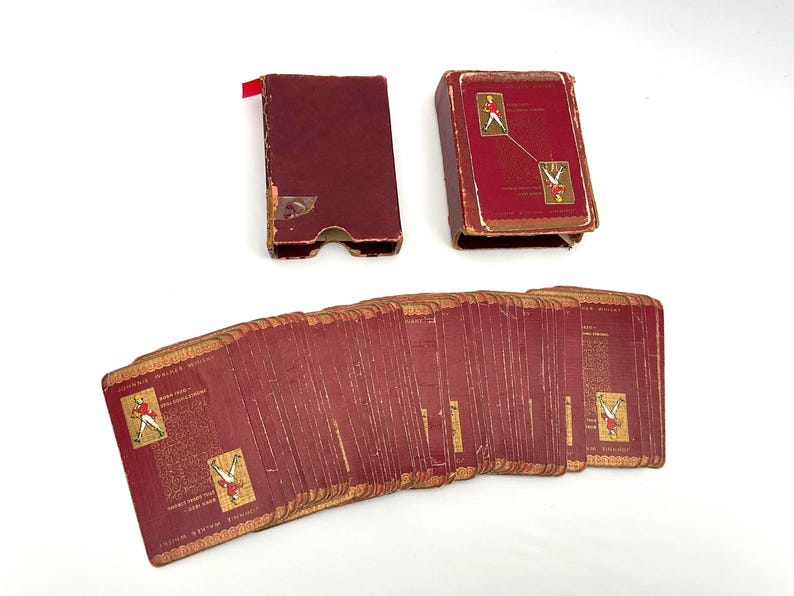 May include: Vintage playing cards with a burgundy and gold design. The deck is fanned out, revealing the card backs and a portion of the faces. The card case is also burgundy with gold accents. The cards feature illustrations of a figure in a red coat.