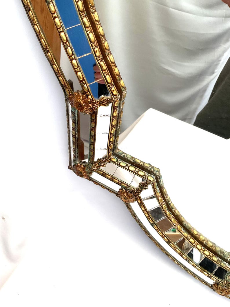 K&ouml;nnte beinhalten: Verzierter, goldgerahmter Spiegel mit einem einzigartigen, geschwungenen Design. Der Rahmen weist aufwendige Details und kleine, quadratische Spiegelfliesen auf. Ein Abschnitt des Spiegels hat blaue Fliesen. Ein dekorativer Einrichtungsgegenstand.