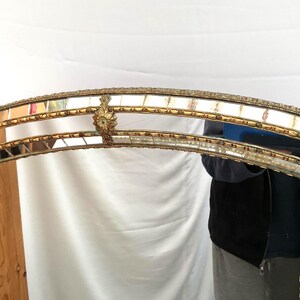K&ouml;nnte beinhalten: Ein gew&ouml;lbter Spiegelrahmen mit einem goldfarbenen, verzierten Design. Der Rahmen besteht aus rechteckigen Spiegelfliesen und dekorativen goldenen Akzenten. Der obere Rand weist eine detaillierte florale Verzierung auf. Der Gesamtstil ist Vintage.