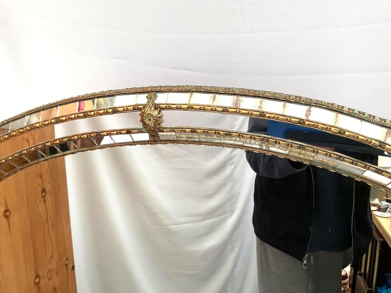 K&ouml;nnte beinhalten: Ein gew&ouml;lbter Spiegelrahmen mit einem goldfarbenen, verzierten Design. Der Rahmen besteht aus rechteckigen Spiegelfliesen und dekorativen goldenen Akzenten. Der obere Rand weist eine detaillierte florale Verzierung auf. Der Gesamtstil ist Vintage.