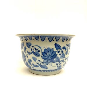 May include: A white ceramic planter with a blue floral design. The planter features a blue lotus flower and leaf pattern. The rim has a wavy blue border. The planter is suitable for indoor or outdoor use.