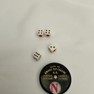 May include: Four white dice with black dots and red pips are scattered on a white surface. A black circular game piece with gold text and the letter "N" is also present. The text on the game piece reads "KWONG FAT CHEUNG H.K."