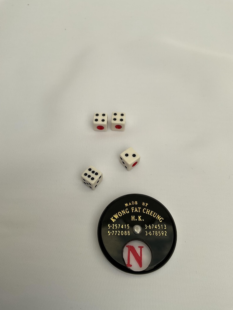 May include: Four white dice with black dots and red pips are scattered on a white surface. A black circular game piece with gold text and the letter "N" is also present. The text on the game piece reads "KWONG FAT CHEUNG H.K."