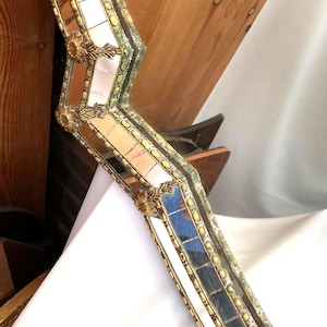 K&ouml;nnte beinhalten: Nahaufnahme eines dekorativen Spiegelrahmens mit Mosaikdesign. Der Rahmen verf&uuml;gt &uuml;ber kleine, rechteckige Spiegelfliesen, goldfarbene Akzente und verzierte florale Eckdetails. Der Rahmen hat eine Vintage- und Antik-&Auml;sthetik.