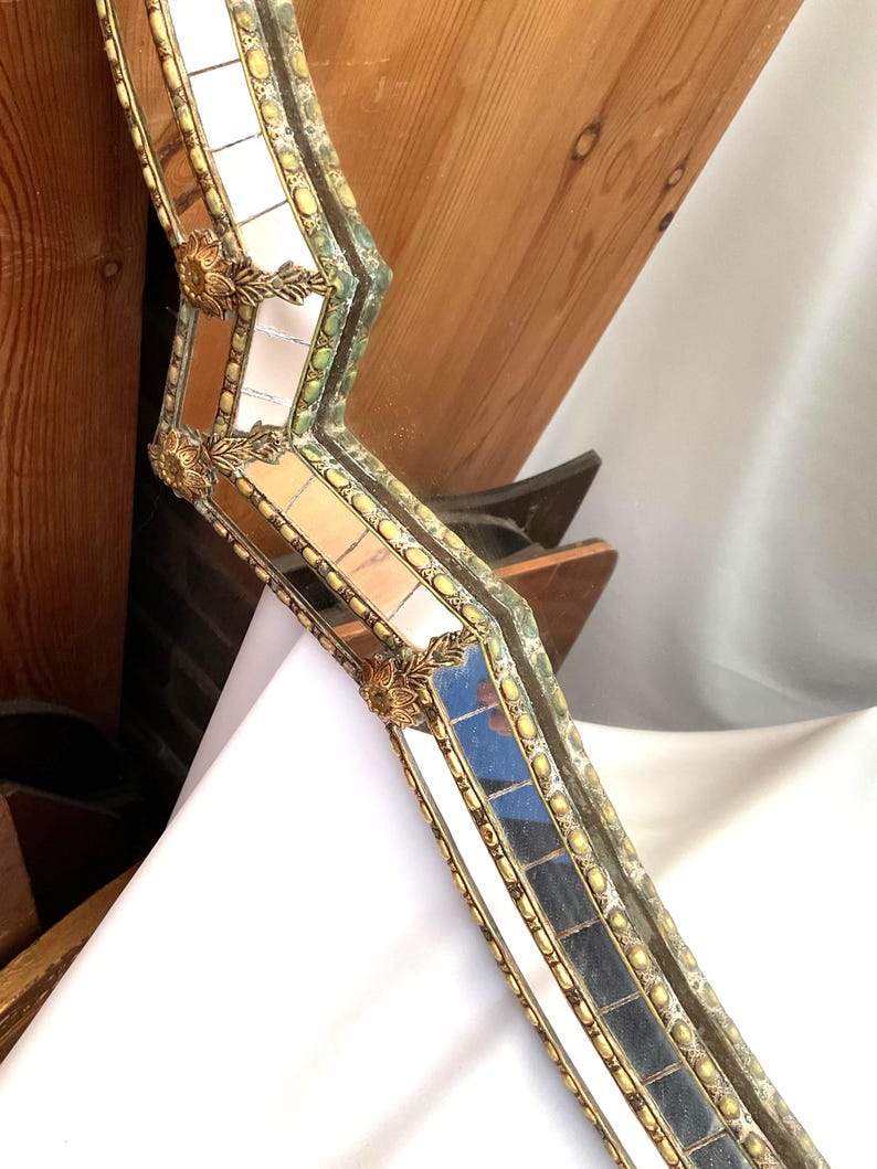 K&ouml;nnte beinhalten: Nahaufnahme eines dekorativen Spiegelrahmens mit Mosaikdesign. Der Rahmen verf&uuml;gt &uuml;ber kleine, rechteckige Spiegelfliesen, goldfarbene Akzente und verzierte florale Eckdetails. Der Rahmen hat eine Vintage- und Antik-&Auml;sthetik.