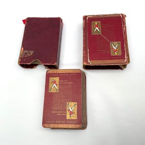 May include: Three vintage Johnnie Walker whisky playing card boxes. The boxes are a deep red with gold accents and feature illustrations of the Johnnie Walker Striding Man. The boxes are open, revealing the cards.