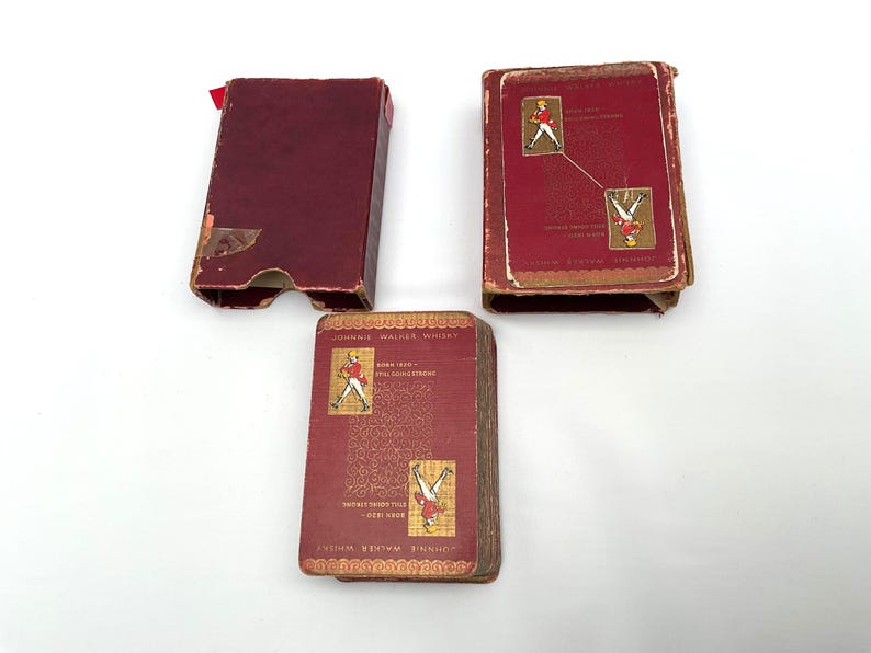 May include: Three vintage Johnnie Walker whisky playing card boxes. The boxes are a deep red with gold accents and feature illustrations of the Johnnie Walker Striding Man. The boxes are open, revealing the cards.