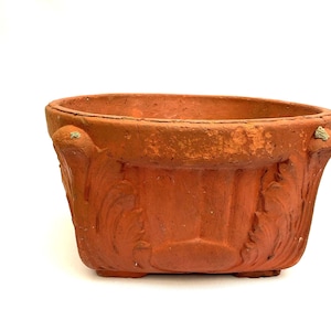 May include: A weathered, terracotta-colored planter with a rectangular shape and ornate, carved floral designs. The planter has small feet and handles on each side. The aged appearance suggests it is an antique or vintage piece, suitable for indoor or outdoor use.