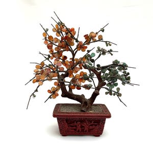 Puede incluir: Un bonsái decorativo con tronco y ramas de color marrón oscuro. Las hojas son de piedras naranjas y verdes. El árbol está en una maceta roja rectangular con figuras talladas. Un artículo de decoración único.