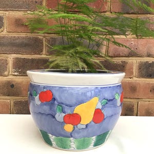 May include: A blue ceramic plant pot with a white rim and a painted fruit design. The pot features red apples, a yellow pear, and green leaves. The pot is filled with a green plant.