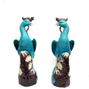 Può includere: Coppia di statue di pavone in ceramica. Le statue presentano corpi turchesi, basi marrone scuro con accenti floreali bianchi e creste marrone scuro. I pavoni si fronteggiano, creando un'esposizione simmetrica.