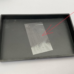 May include: A black rectangular tray with a textured surface. A clear, rectangular piece of plastic tape is placed inside the tray. The tray has a raised edge.