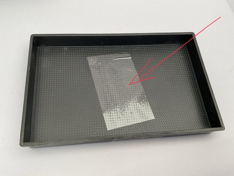 May include: A black rectangular tray with a textured surface. A clear, rectangular piece of plastic tape is placed inside the tray. The tray has a raised edge.