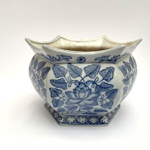 Può includere: Un vaso da fiori esagonale in ceramica con base bianca e motivi floreali e di foglie blu. Il bordo presenta una forma smerlata e la base è decorata con un motivo geometrico a rombi. L'interno è bianco semplice e l'esterno è smaltato.