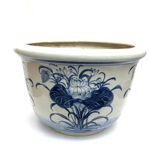 May include: A white ceramic planter with a blue floral pattern. The planter has a wide, rounded form with a gently flared rim. The blue design features lotus flowers and leaves, adding a touch of elegance to the piece.