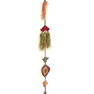 Può includere: Un ornamento decorativo da appendere in verticale caratterizzato da un lungo cordino decorato con diversi elementi, tra cui una nappa color pesca, un componente in tessuto rosso con due nappe verde oliva, un piccolo ciondolo grigio e una nappa rossa.