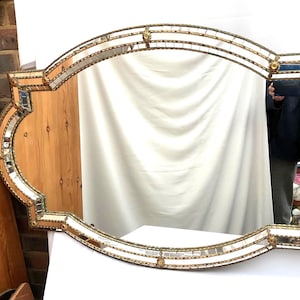 K&ouml;nnte beinhalten: Ein verzierter Spiegel im antiken Stil mit einem goldfarbenen Rahmen. Der Spiegel hat eine ovale Form mit dekorativen Kanten und kleinen Spiegelfliesen. Der Rahmen hat ein detailliertes, strukturiertes Design, das seinen Vintage-Reiz unterstreicht.