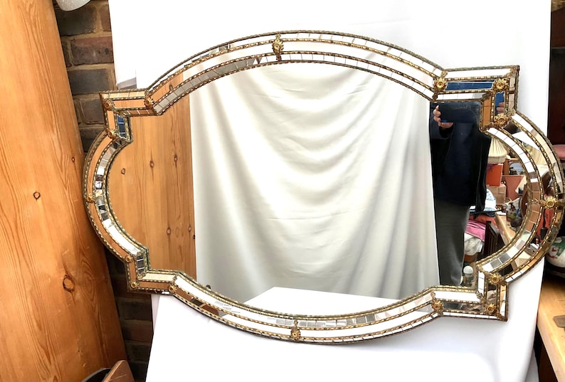 K&ouml;nnte beinhalten: Ein verzierter Spiegel im antiken Stil mit einem goldfarbenen Rahmen. Der Spiegel hat eine ovale Form mit dekorativen Kanten und kleinen Spiegelfliesen. Der Rahmen hat ein detailliertes, strukturiertes Design, das seinen Vintage-Reiz unterstreicht.