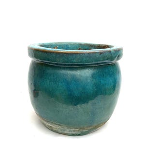 May include: A small, round, teal-colored ceramic pot with a wide rim. The pot has a glossy finish and a slightly textured surface. The color varies from a deep teal to a lighter turquoise, with some areas showing a hint of brown. Ideal for small plants.