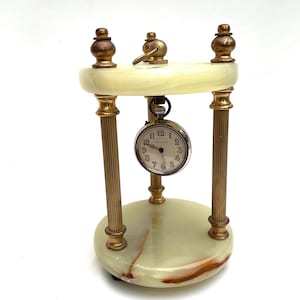 May include: A vintage desk clock featuring a cream onyx base and top, connected by gold-toned metal columns. A small, round clock with a white face and black numerals is suspended in the centre. The clock has a key for winding.