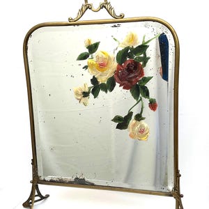May include: An antique mirror with a brass frame and stand. The mirror features a floral painting of yellow and burgundy roses with green leaves. The frame has a decorative handle.