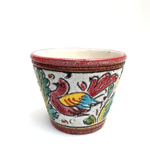 May include: A small, ceramic planter with a colorful bird and floral design. The planter features a white base with a red rim and a brown textured band. The bird is depicted with red, yellow, and blue feathers.