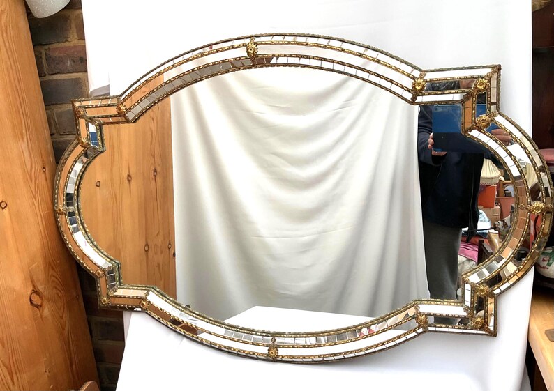 K&ouml;nnte beinhalten: Ein verzierter, gro&szlig;er, rechteckiger Spiegel mit einem dekorativen goldfarbenen Rahmen. Der Rahmen ist mit kleinen Spiegelfliesen und goldenen Akzenten verziert. Die Form des Spiegels ist ein einzigartiges, geschwungenes Design, geeignet f&uuml;r Wanddekoration.