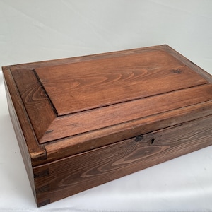 May include: A dark brown wooden box with a hinged lid and a keyhole. The box has a simple design with a raised edge around the lid.