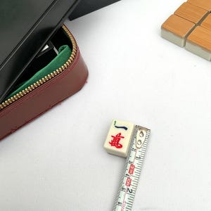 May include: A close-up of a Mahjong tile with red and blue characters, measuring approximately 2.5 cm. The tile is cream-colored with a black case and a red leather zipper case with a green interior. Additional tiles are visible.