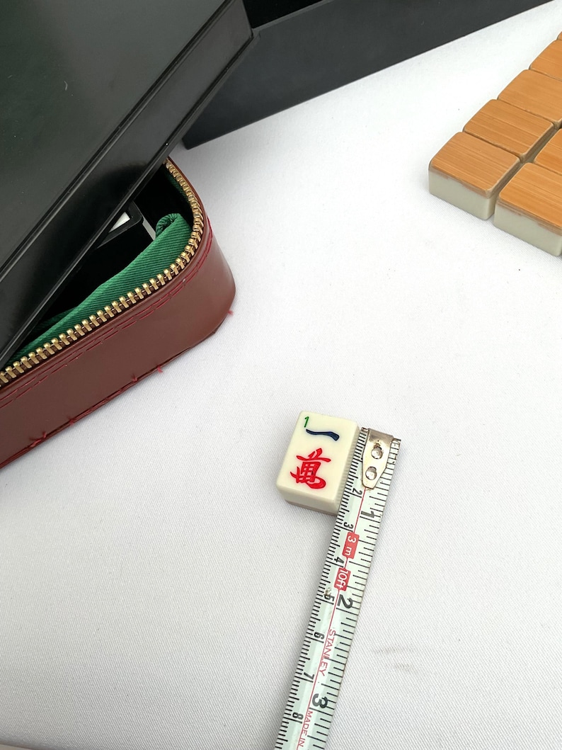 May include: A close-up of a Mahjong tile with red and blue characters, measuring approximately 2.5 cm. The tile is cream-colored with a black case and a red leather zipper case with a green interior. Additional tiles are visible.