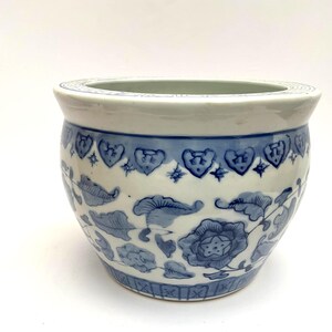 May include: A round ceramic planter featuring a white base with blue hand-painted floral patterns and leaf motifs. The rim is decorated with a repeating heart-shaped border, while the base displays a geometric grid pattern in blue and white.