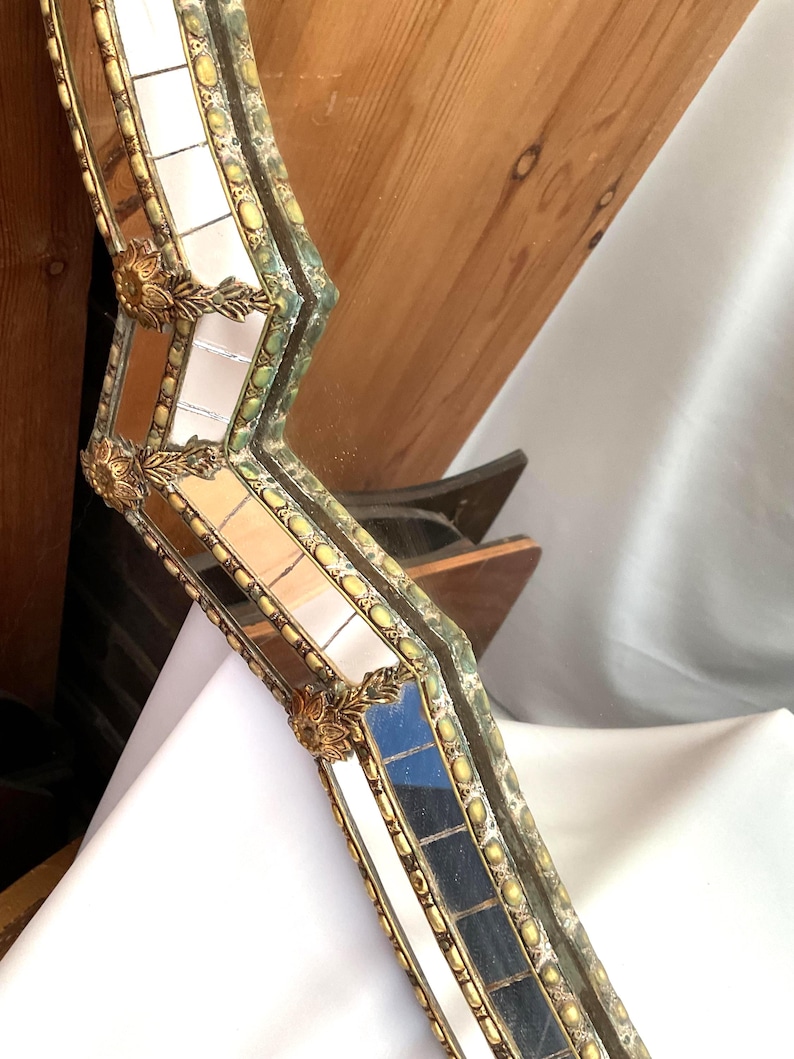 K&ouml;nnte beinhalten: Ein verzierter, antiker Spiegelrahmen mit goldener und gr&uuml;ner Oberfl&auml;che. Der Rahmen ist mit kleinen Spiegelfliesen und dekorativen goldenen Akzenten segmentiert. Der Rahmen hat eine einzigartige, eckige Form und ist vor einem neutralen Hintergrund platziert.