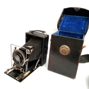 May include: A vintage black camera with a collapsible bellows and a silver lens, next to its matching black leather case. The case has a blue velvet interior and a brass lock. A black strap is attached to the case.