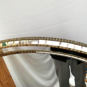 K&ouml;nnte beinhalten: Ein dekorativer Spiegel mit einem goldfarbenen Rahmen und einem Mosaik-Bord. Der Rahmen hat verzierte Details und eine geschwungene Form. Die Oberfl&auml;che des Spiegels ist reflektierend, und das Gesamtdesign ist von Vintage inspiriert.