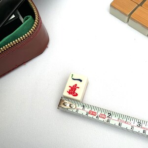 May include: A Mahjong tile showing the number one and Chinese characters in red and blue, being measured with a tape measure. The tile is approximately 2.5 cm long. A burgundy case with a gold zipper is in the background.