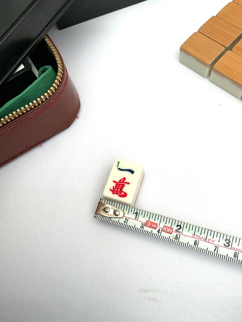 May include: A Mahjong tile showing the number one and Chinese characters in red and blue, being measured with a tape measure. The tile is approximately 2.5 cm long. A burgundy case with a gold zipper is in the background.