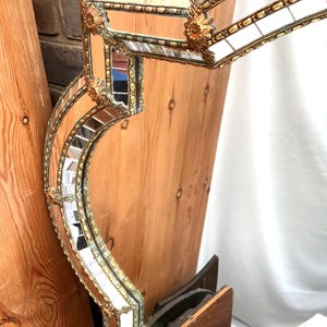 K&ouml;nnte beinhalten: Verzierter Vintage-Spiegel mit goldfarbenem Rahmen und kleinen Spiegelfliesen. Der Rahmen weist dekorative Blumenakzente und ein geschwungenes, geometrisches Design auf. Die reflektierende Oberfl&auml;che und die aufwendigen Details des Spiegels sind sichtbar.