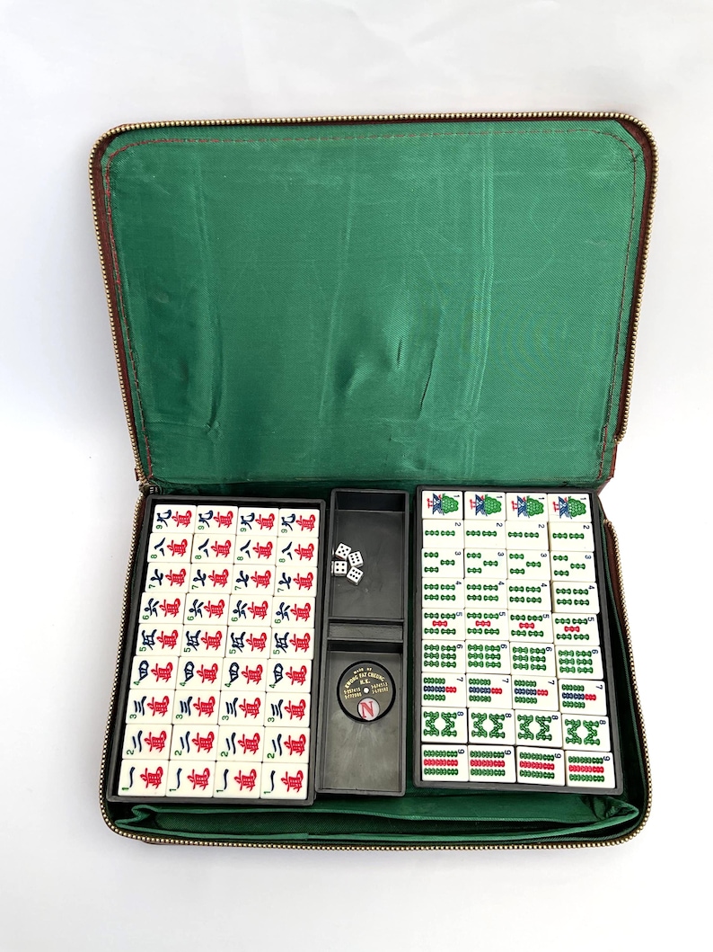 May include: A vintage Mahjong set in a brown zipped case with a green lining. The set contains tiles with red and green symbols, dice, and a circular marker. The tiles are arranged in black trays.