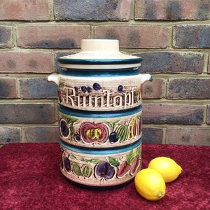May include: A ceramic jar with a lid, decorated with a colourful fruit pattern and the word "Rumtopf". The jar is sitting on a red surface with two yellow lemons in front of it.